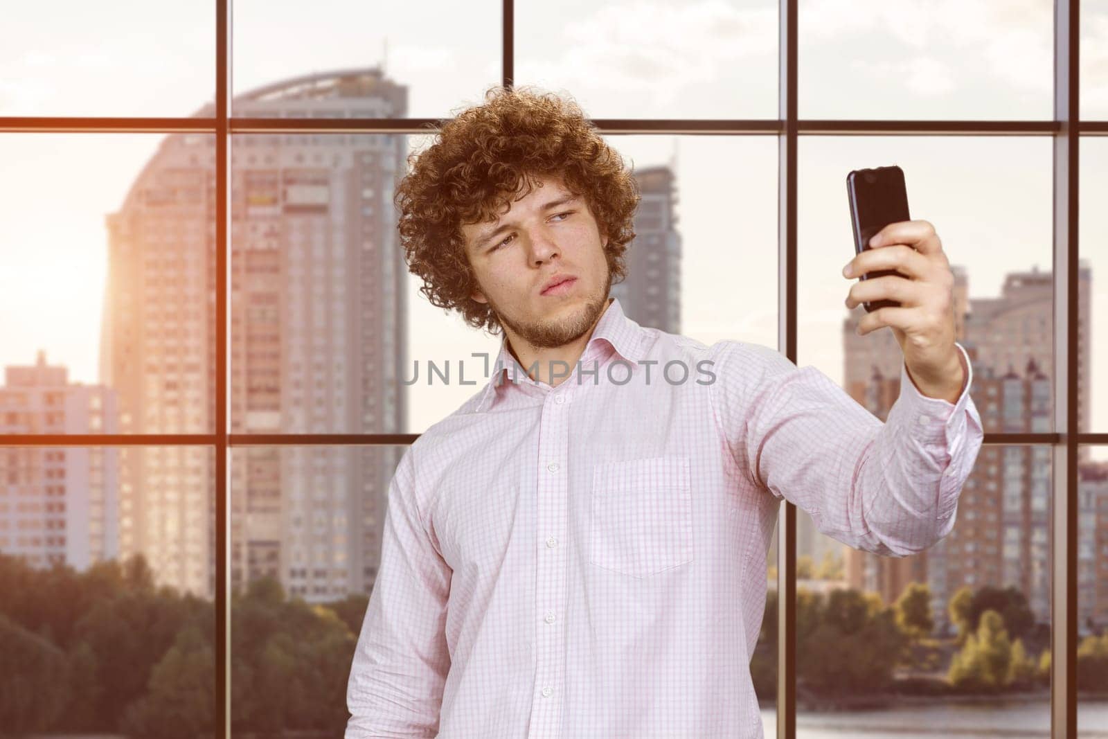 Serious young man with curly hair taking selfie with smartphone. Checkered indoor background.