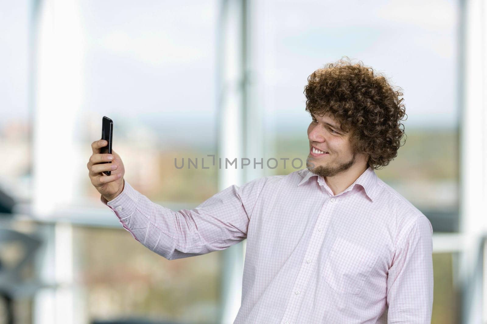 Portrait of happy young man with curly hair taking selfie with smartphone. Indoor window in the background.