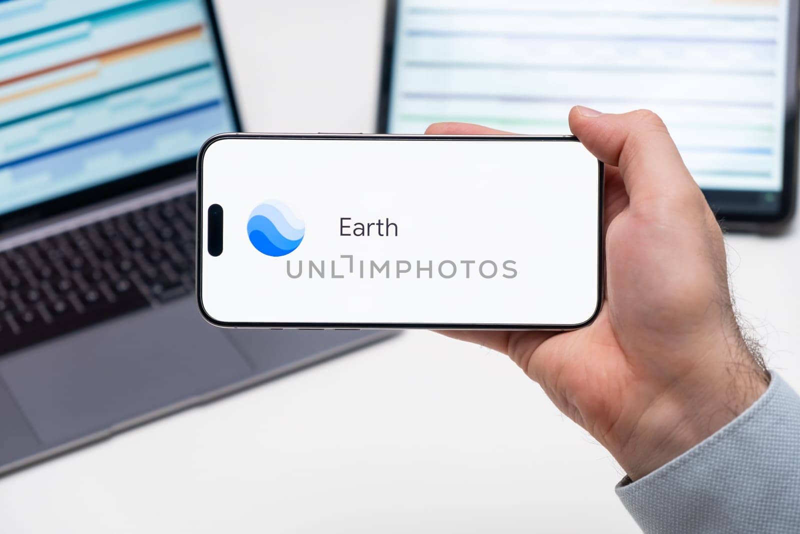 Earth application logo on the screen of smart phone in mans hand, laptop and tablet on the table by vladimka