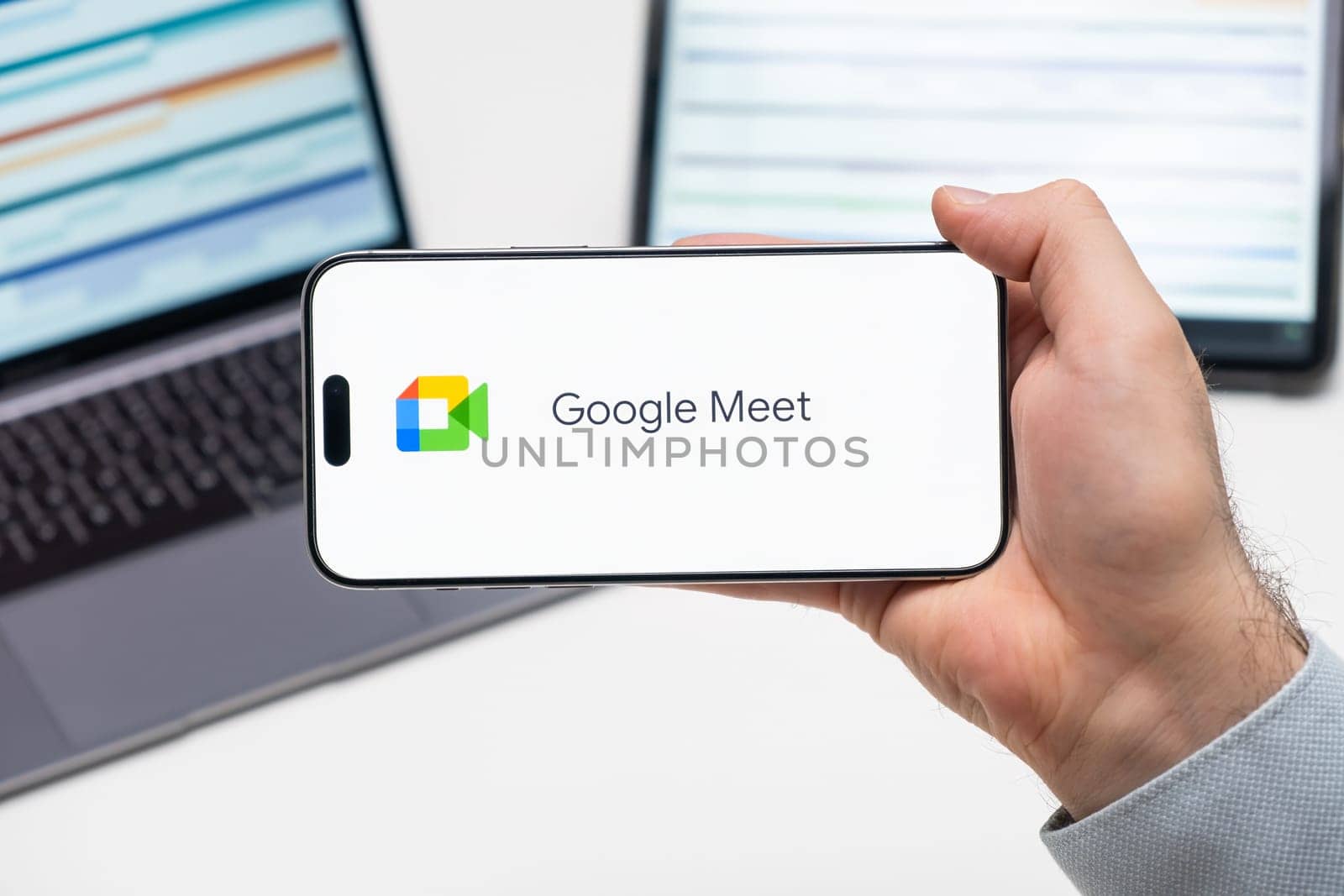Google Meet logo of app on the screen of mobile phone held by man in front of the laptop and tablet, December 2023, Prague, Czech Republic