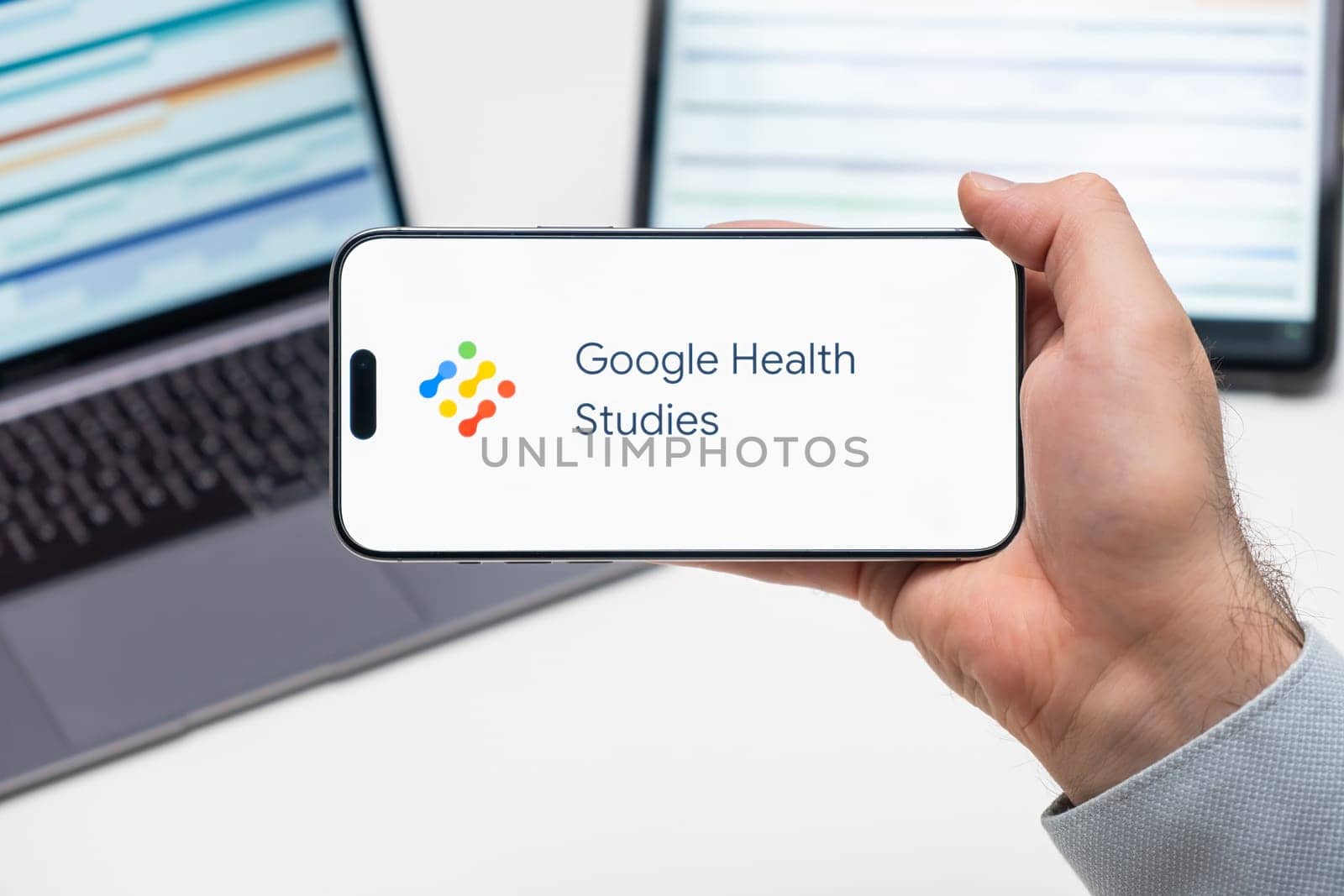 Google Health Studies logo of app on the screen of mobile phone held by man in front of the laptop and tablet, December 2023, Prague, Czech Republic