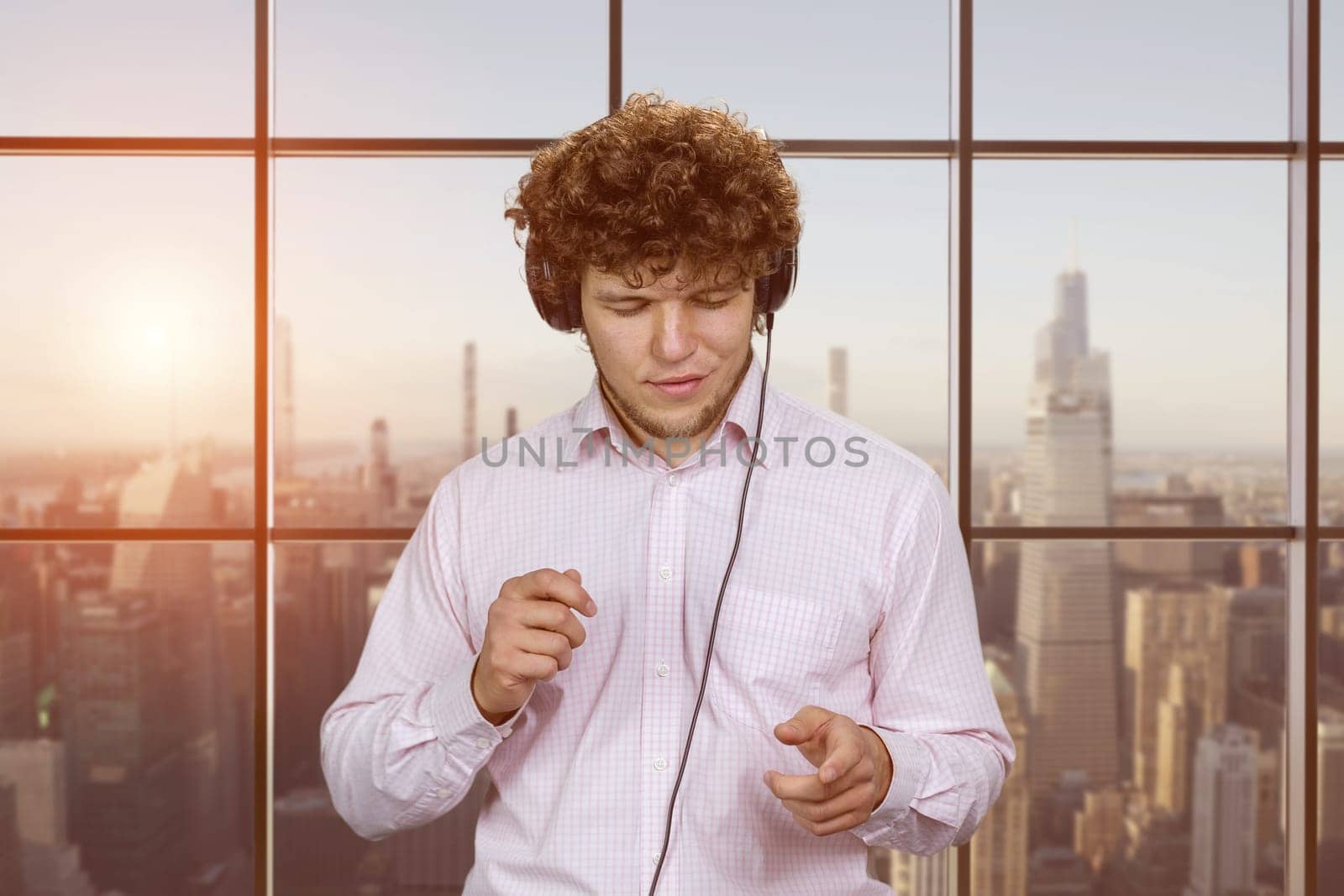 Portrait of happy young guy enjoying music in headphones. Indoor window with cityscape view in the background.