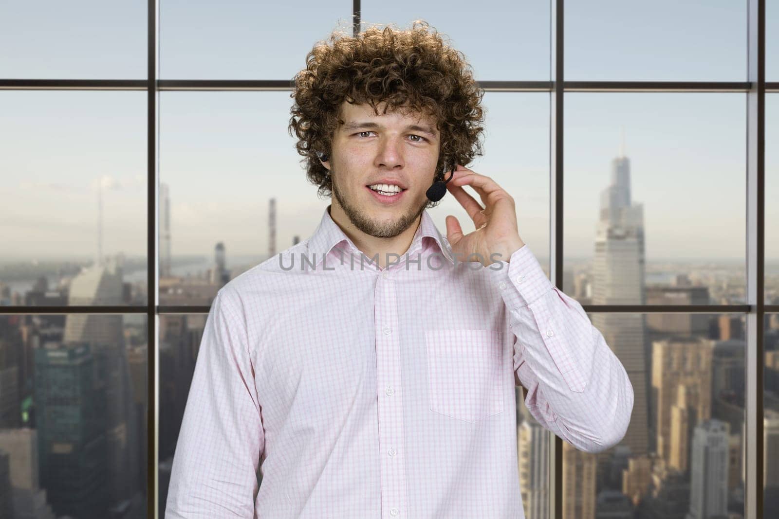 Portrait of a happy young successful man talking in microphone touching in with finger. Checkered window with cityscape view in the background.