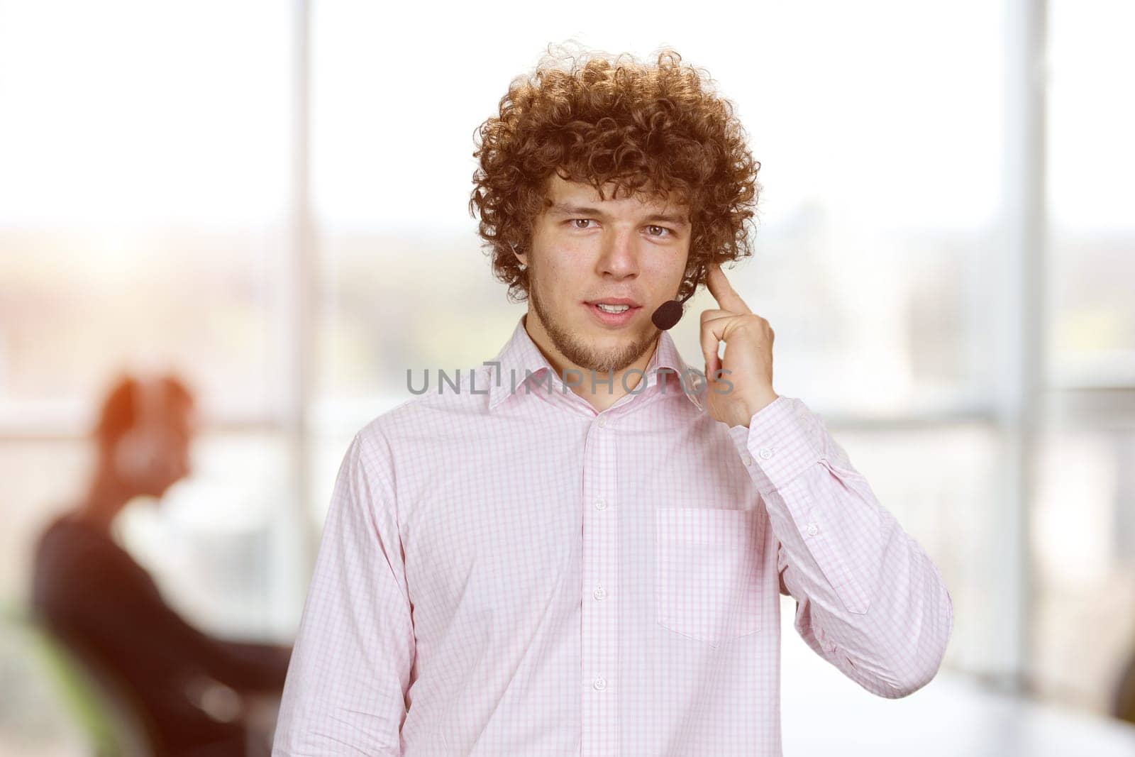 Young serious successful man in curly hair talking in microphone. Office interior in the background.