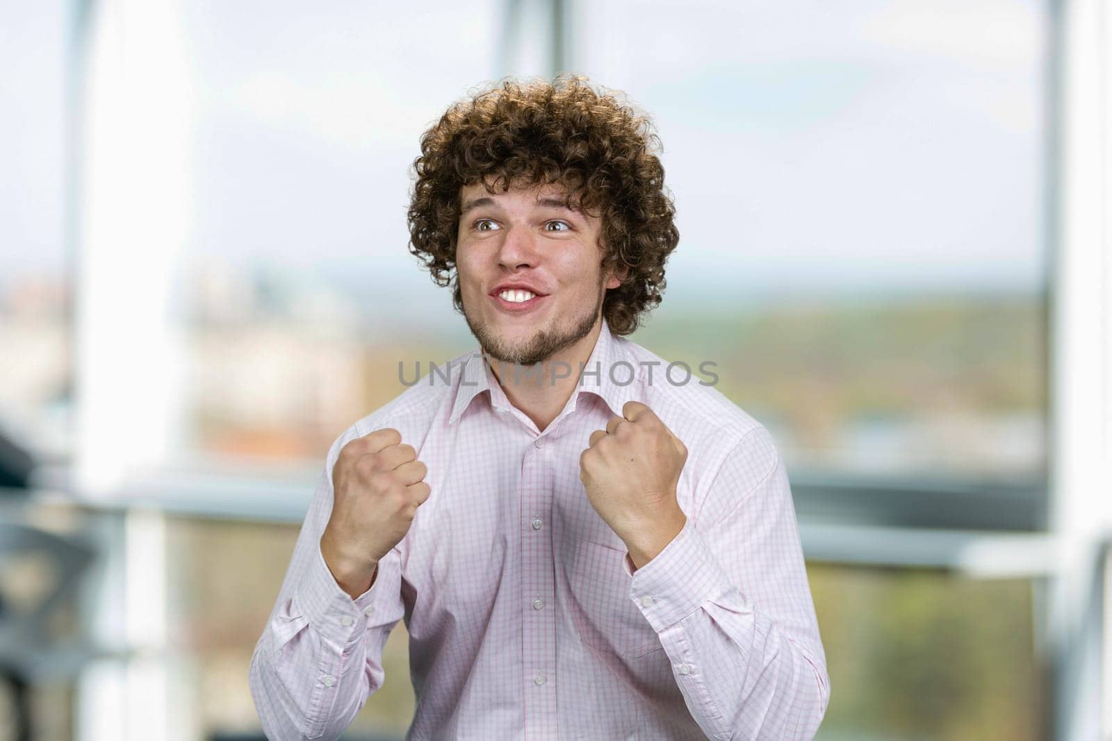 Portrait of happy excited young man with curly hair rejoicing success. Young football fan. Indoor window in the background.