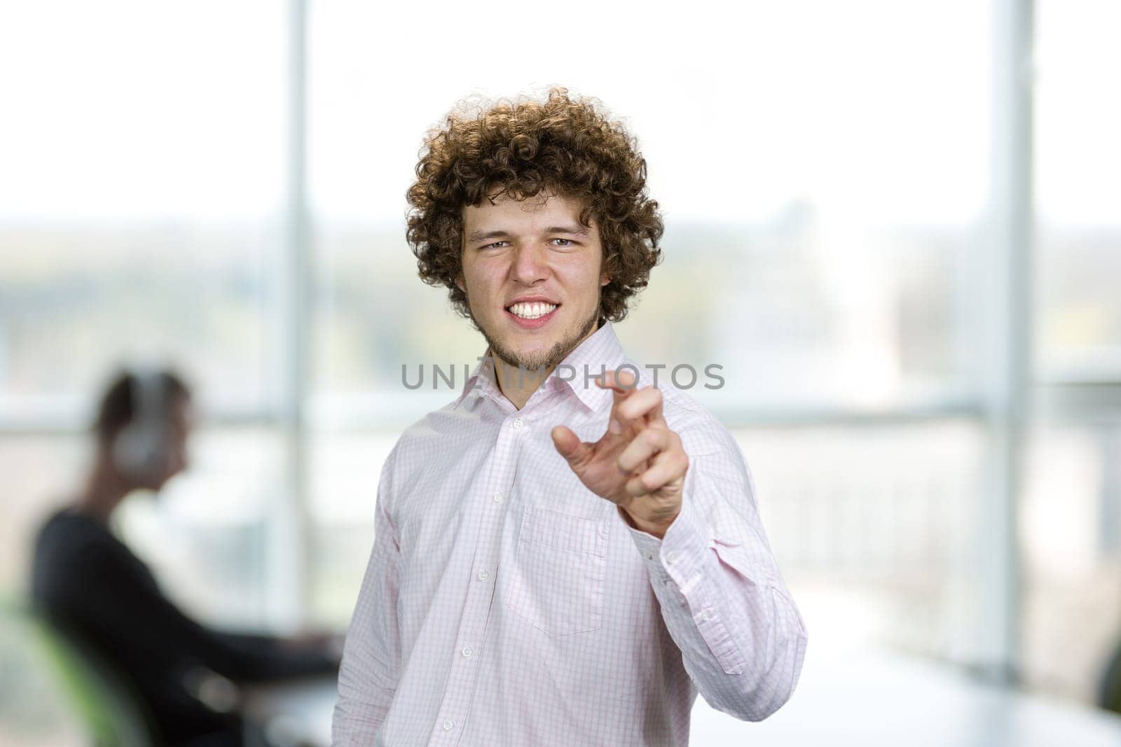 Portrait of a happy young cheerful man with curly hair shows his teeth and gesturing something. Blurred office interior in the background.