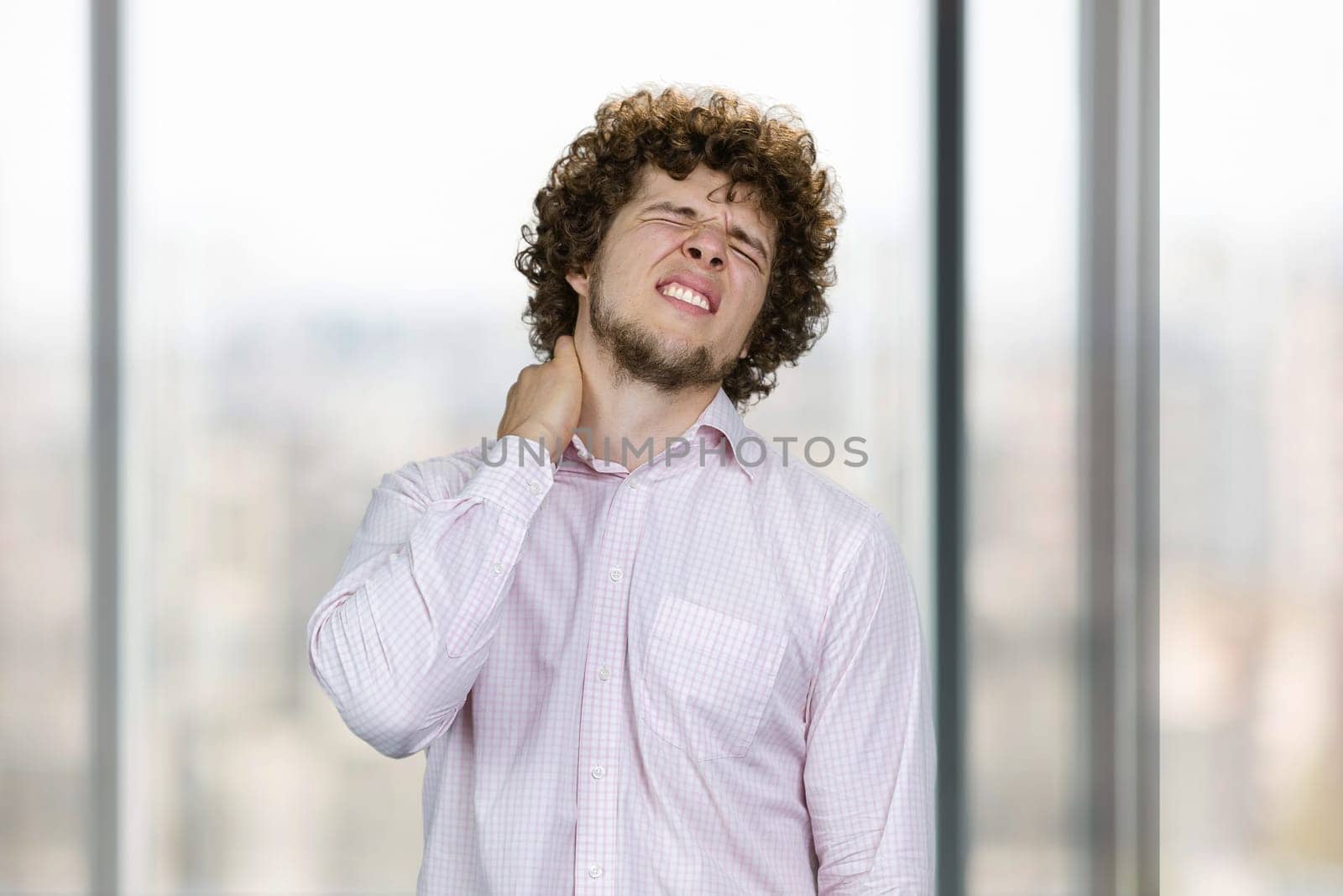 Young handsome man with curly hair having neckache. Indoor window in the background.