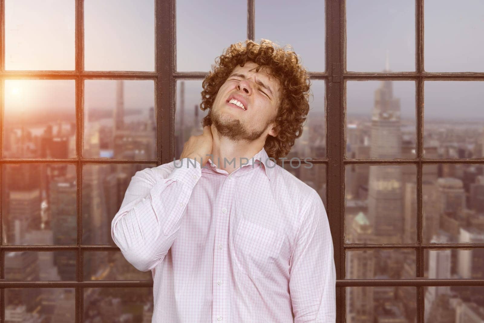 Portrait of a young caucasian man with curly hair having a neckache. Window with cityscape view in the background.