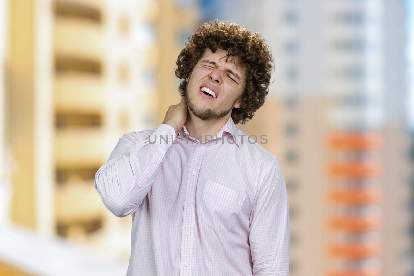 Young handsome man with curly hair having neckache. Residential area in the background.