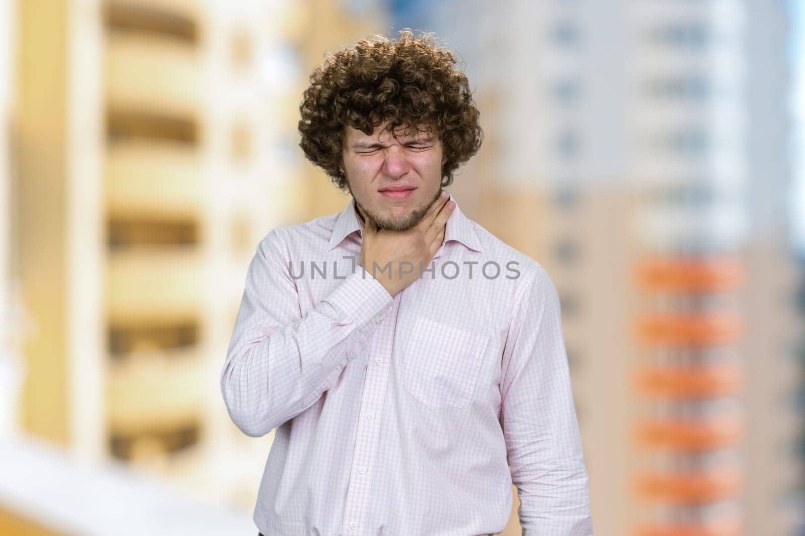 Young handsome man with curly hair touching his neck. Residential area buildings in the background.