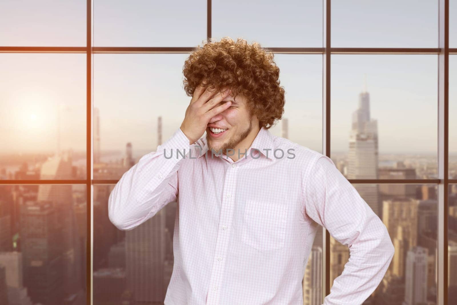 Portrait of a young man with curly hair facepalming because of a cringe. Checkered window with evening cityscape view.