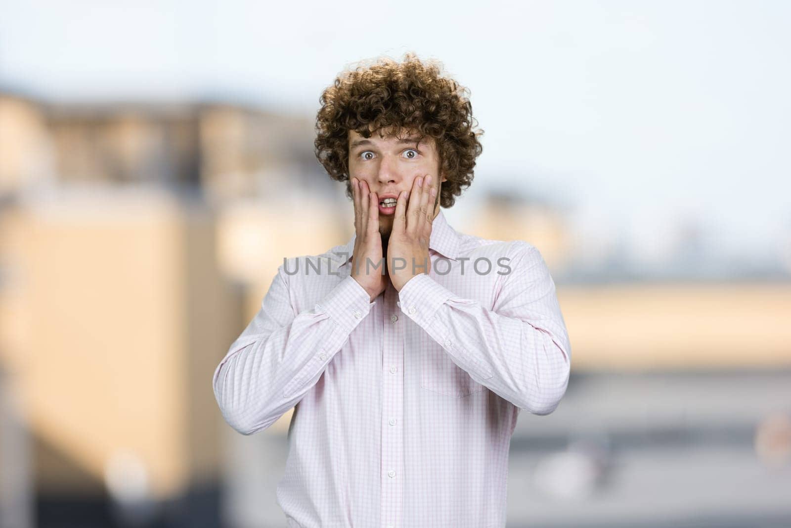 Portrait of shocked amazed young guy with curly hair touching his face. Blurred urban background.