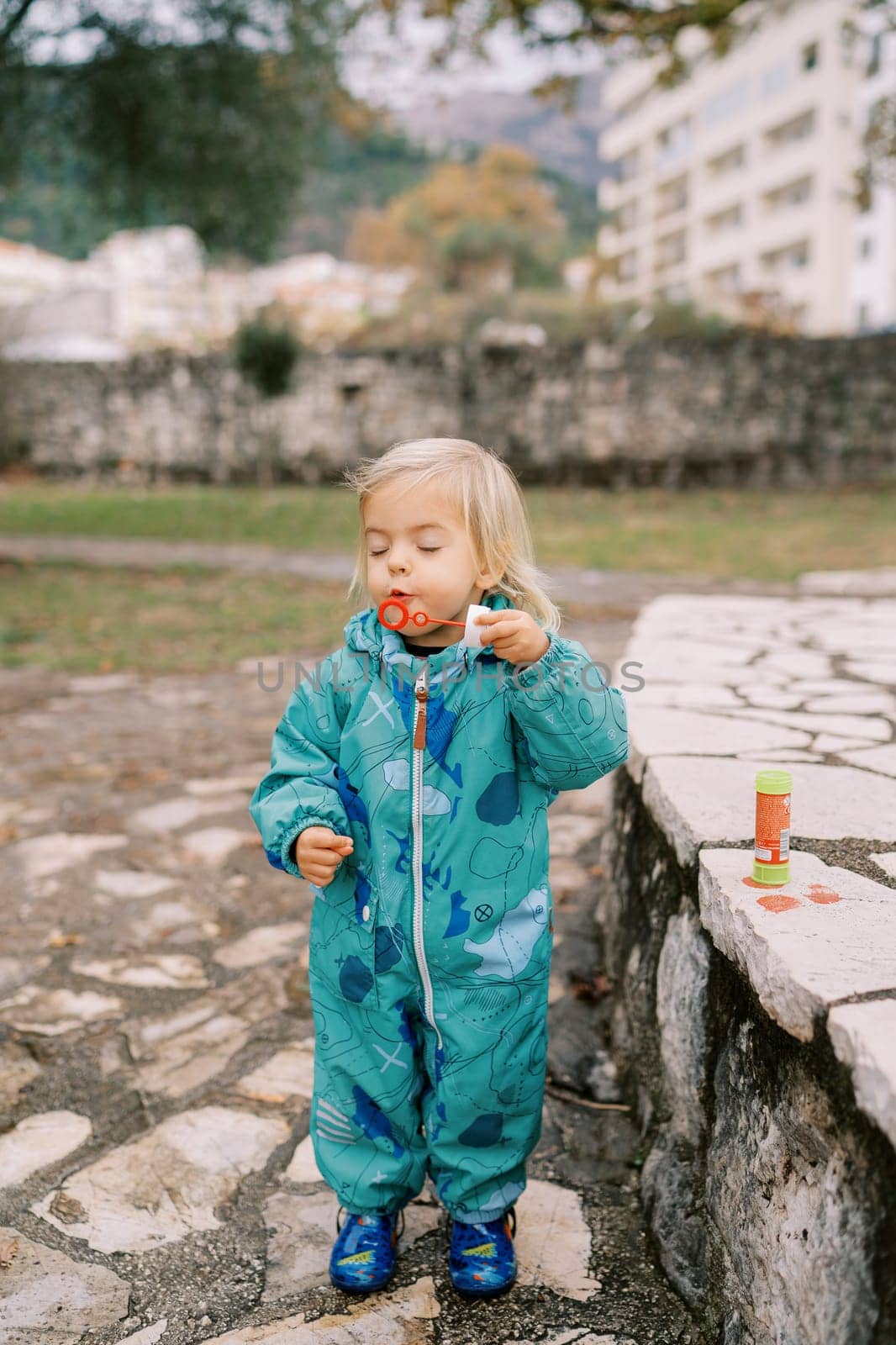 Little girl in overalls blowing soap bubbles squinting her eyes in the park. High quality photo