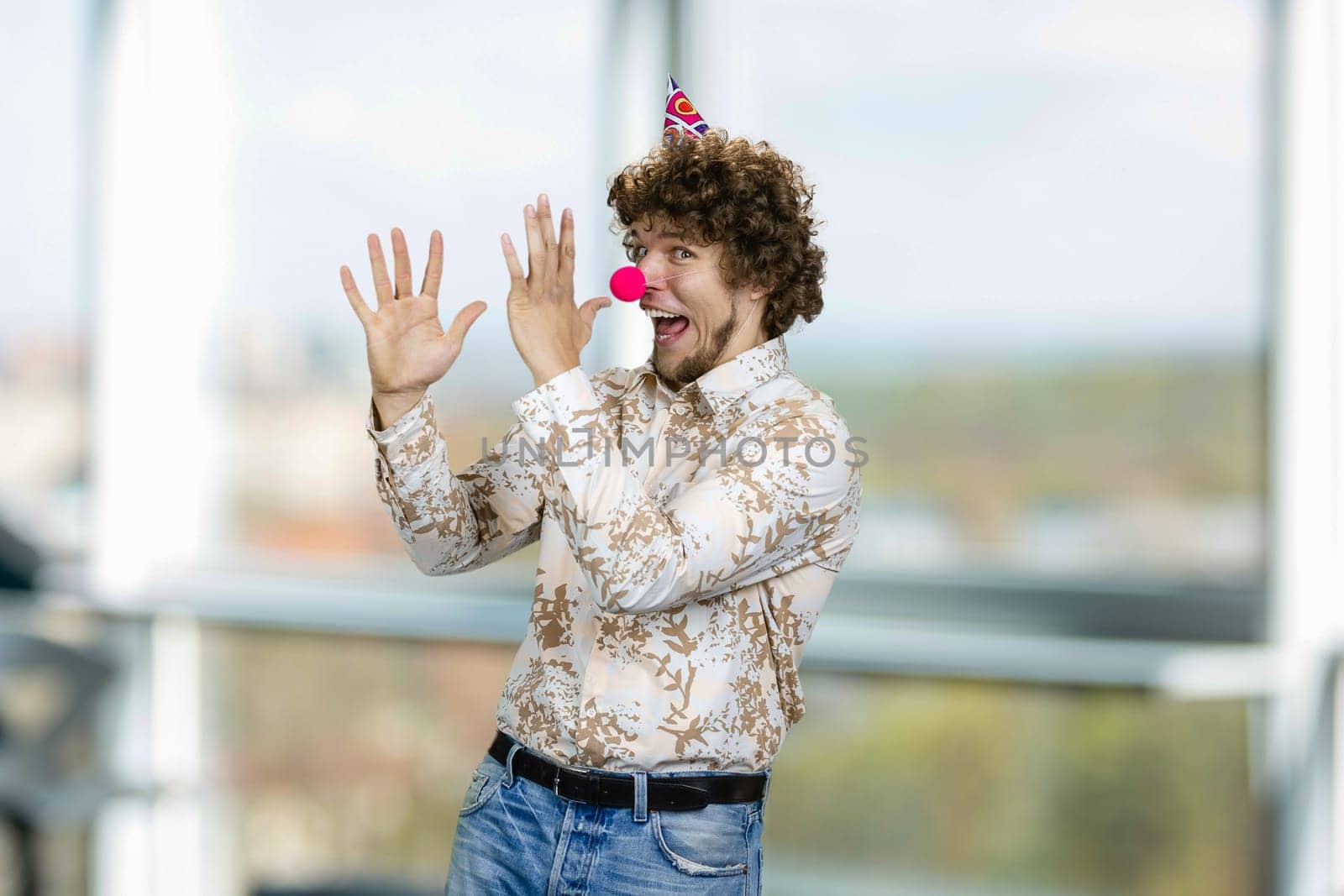 Happy young caucasian guy with curly hair wearing a clowns nose. Indoor window in the background.