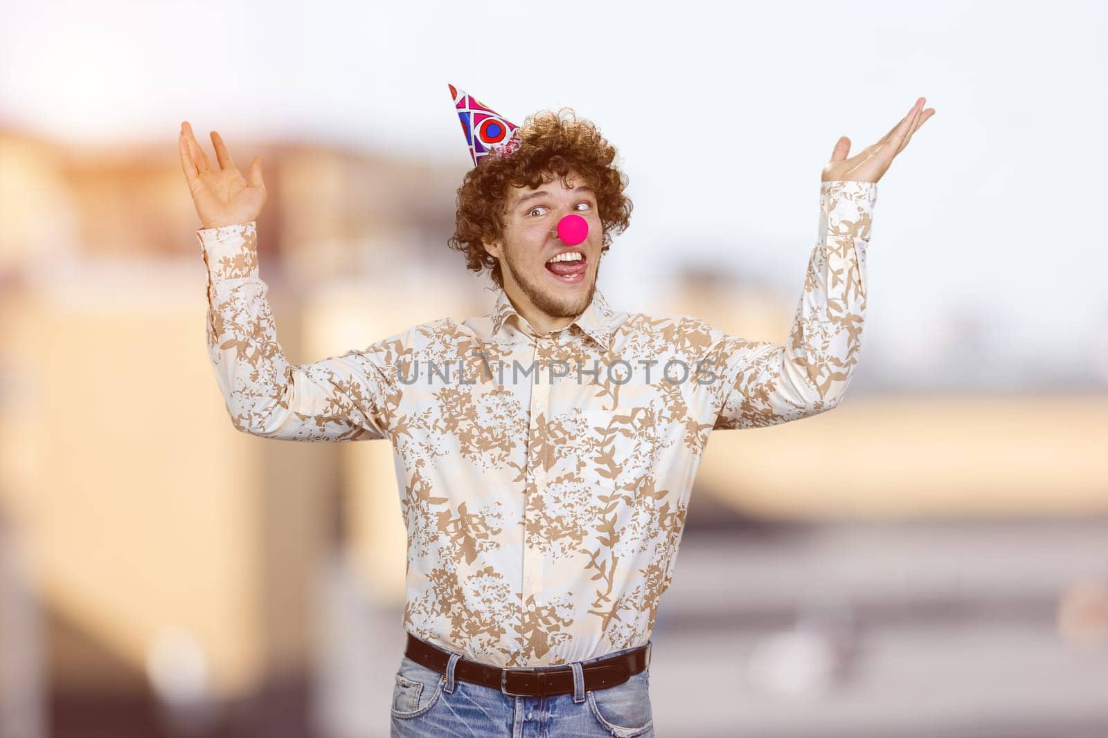 Happy young guy with clown nose fooling around making funny faces. Blurred cityscape in the background.