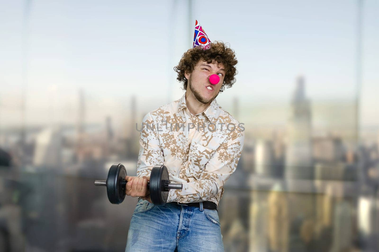 Portrait of a young clown making a gym exercise with dumbbell. Blurred cityscape in the background.