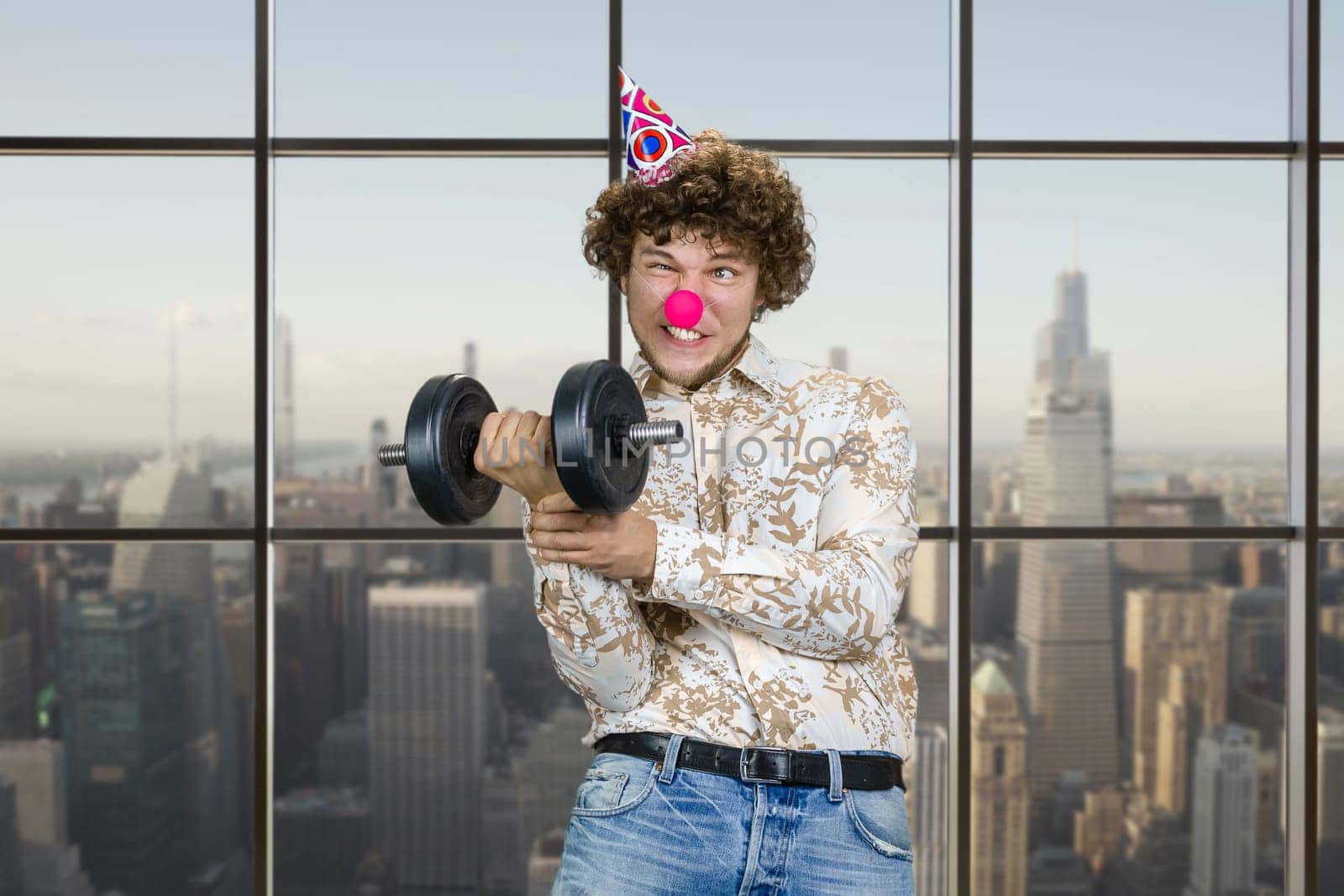 Portrait of a young clown making funny face and holding a dumbbell indoors. Checkered window with cityscape view in the background.