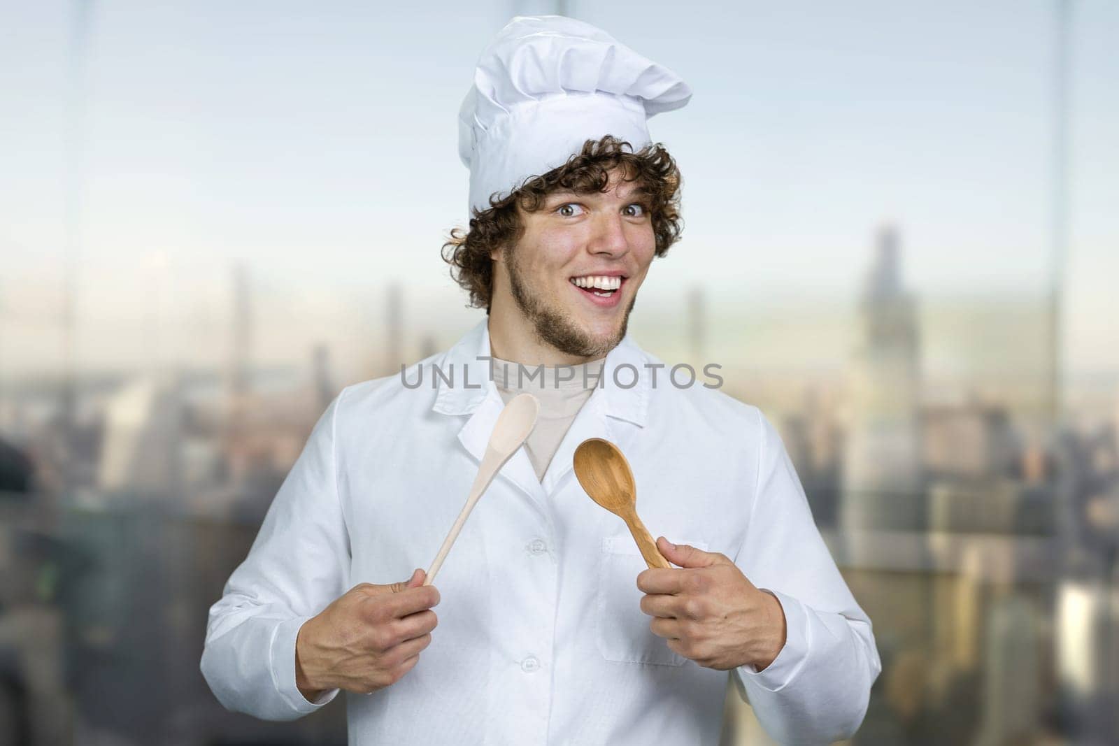 Portrait of a young male cook with a wooden spoon. Cooking food concept. Blurred cityscape in the background.