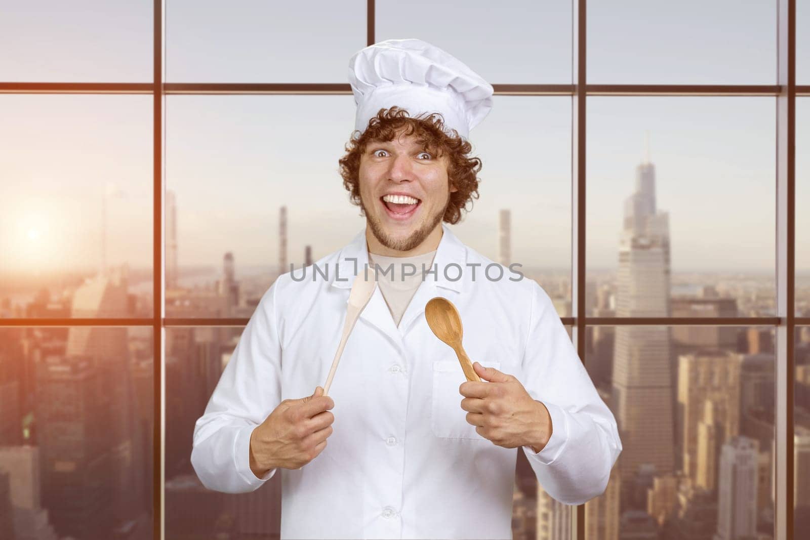 Portrait of a happy young caucasian male cook with a wooden spoon. Checkered window with cityscape in the background.