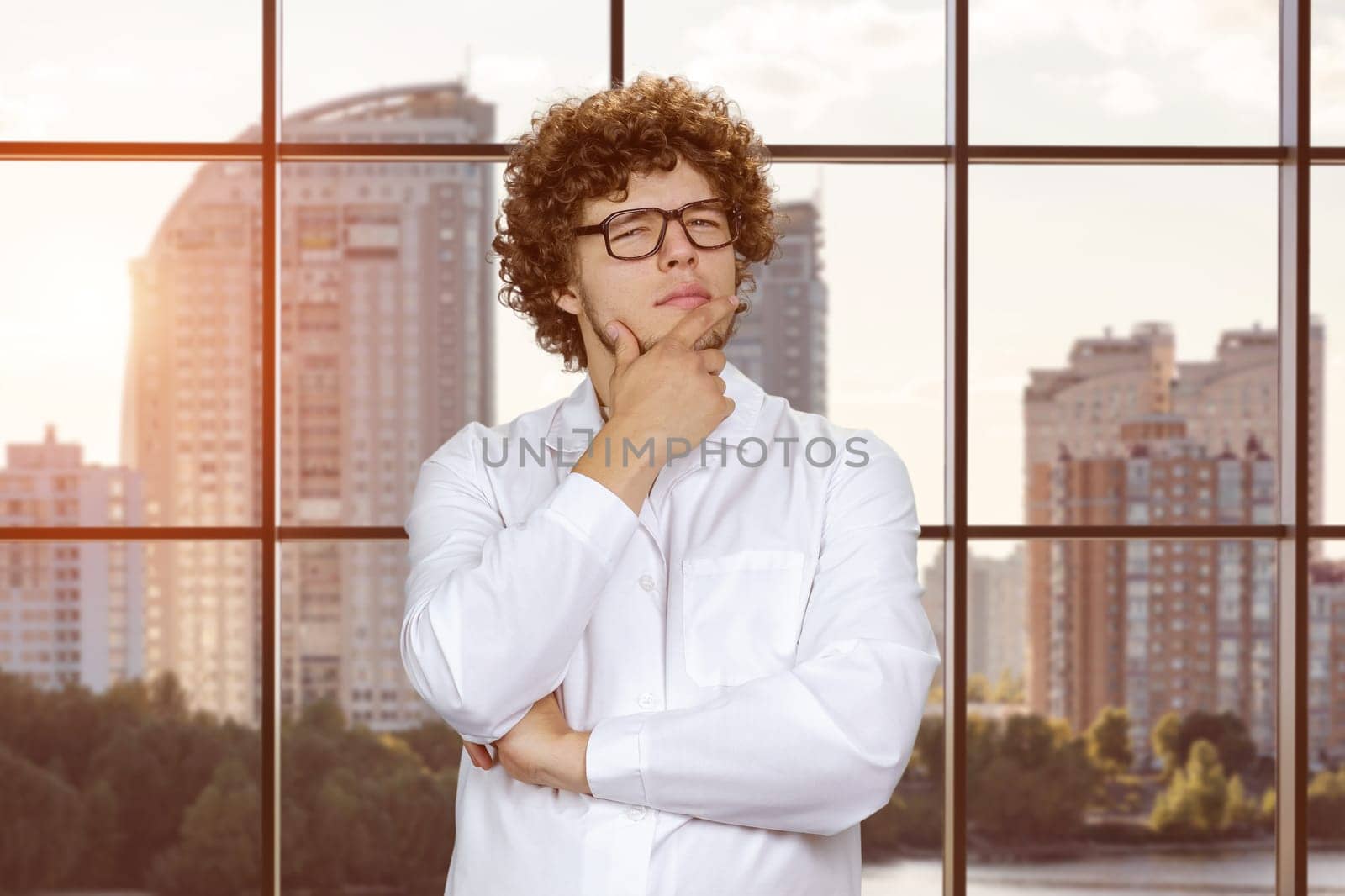 Portrait of a smart young man with glasses thinking about something. Checkered window with cityscape view in the background.