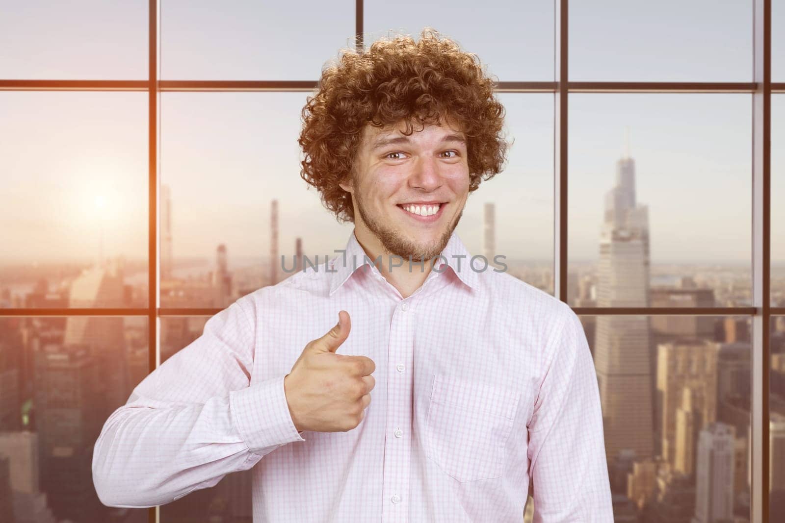 Portrait of a happy young man with curly hair shows thumb up. Checkered window with cityscape view.