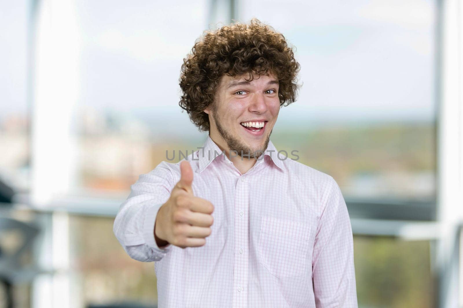 Happy smiling young man with curly hair shows thumb up. Indoor window in the background.