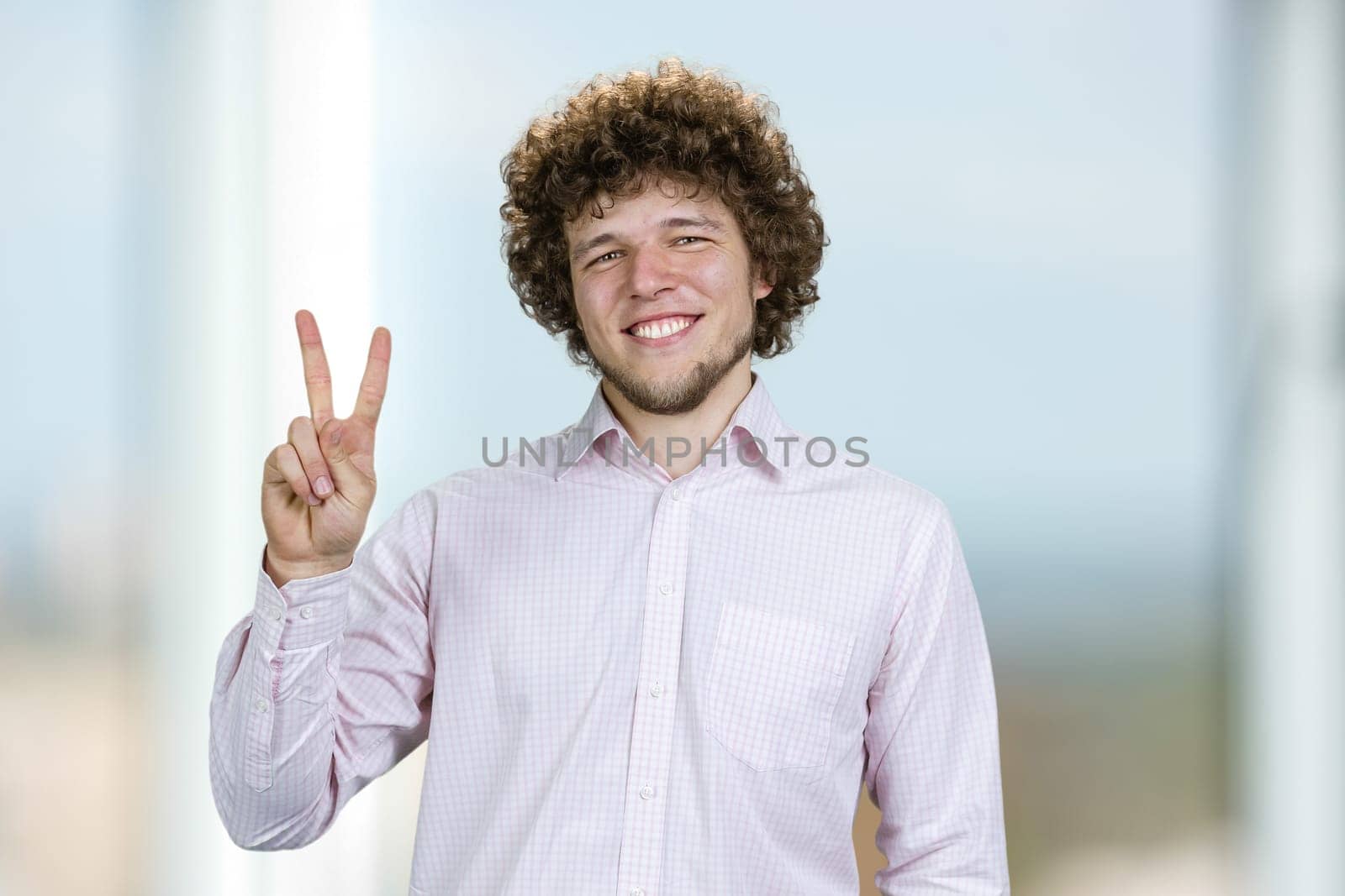 Portrait of a happy young man with curly hair shows victory gesture sign. Indoor window in the background.