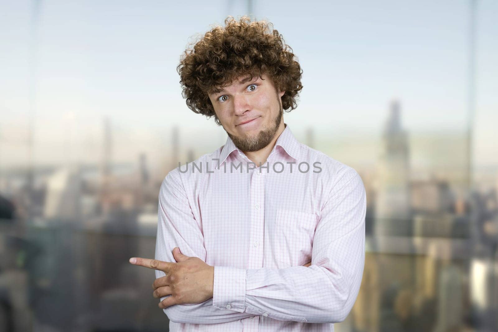 Portrait of a young man with curly hair pointing to the left with his index finger. Blurred cityscape in the background.