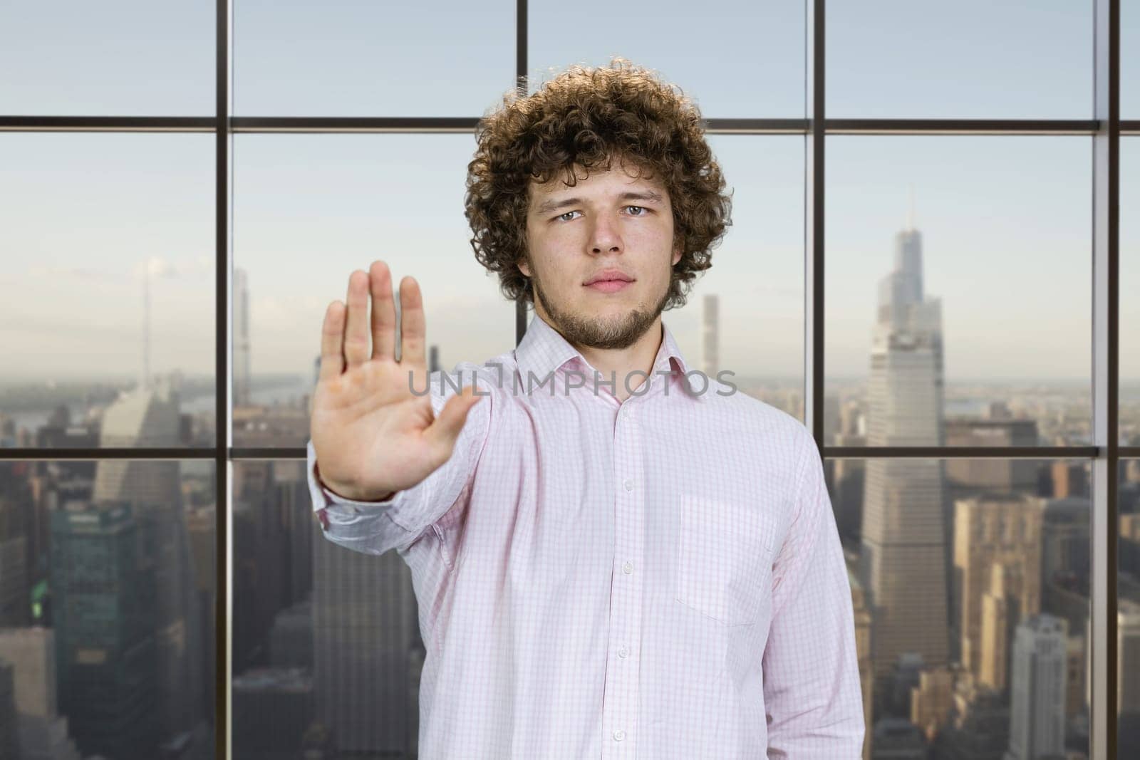 Serious young man with curly hair shows his stop gesture sign. Checkered window with cityscape view in the background.