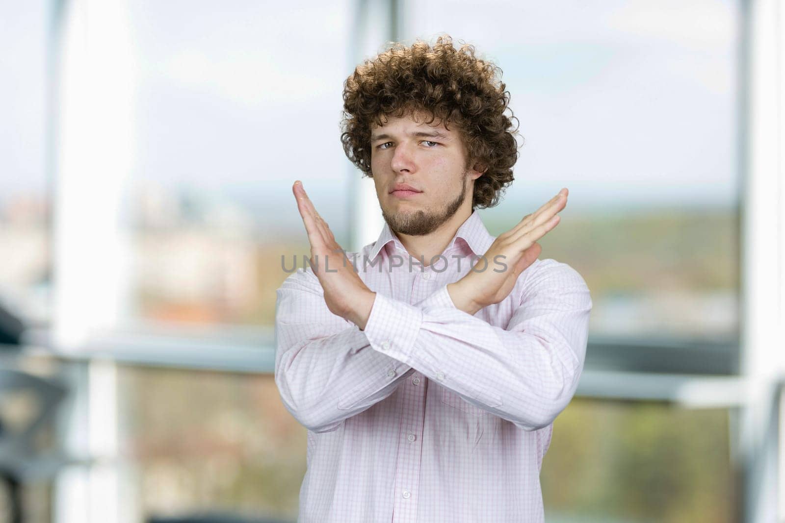 Portrait of a young man with curly hair cross his arms as sign of prohibition. Office window in the background.
