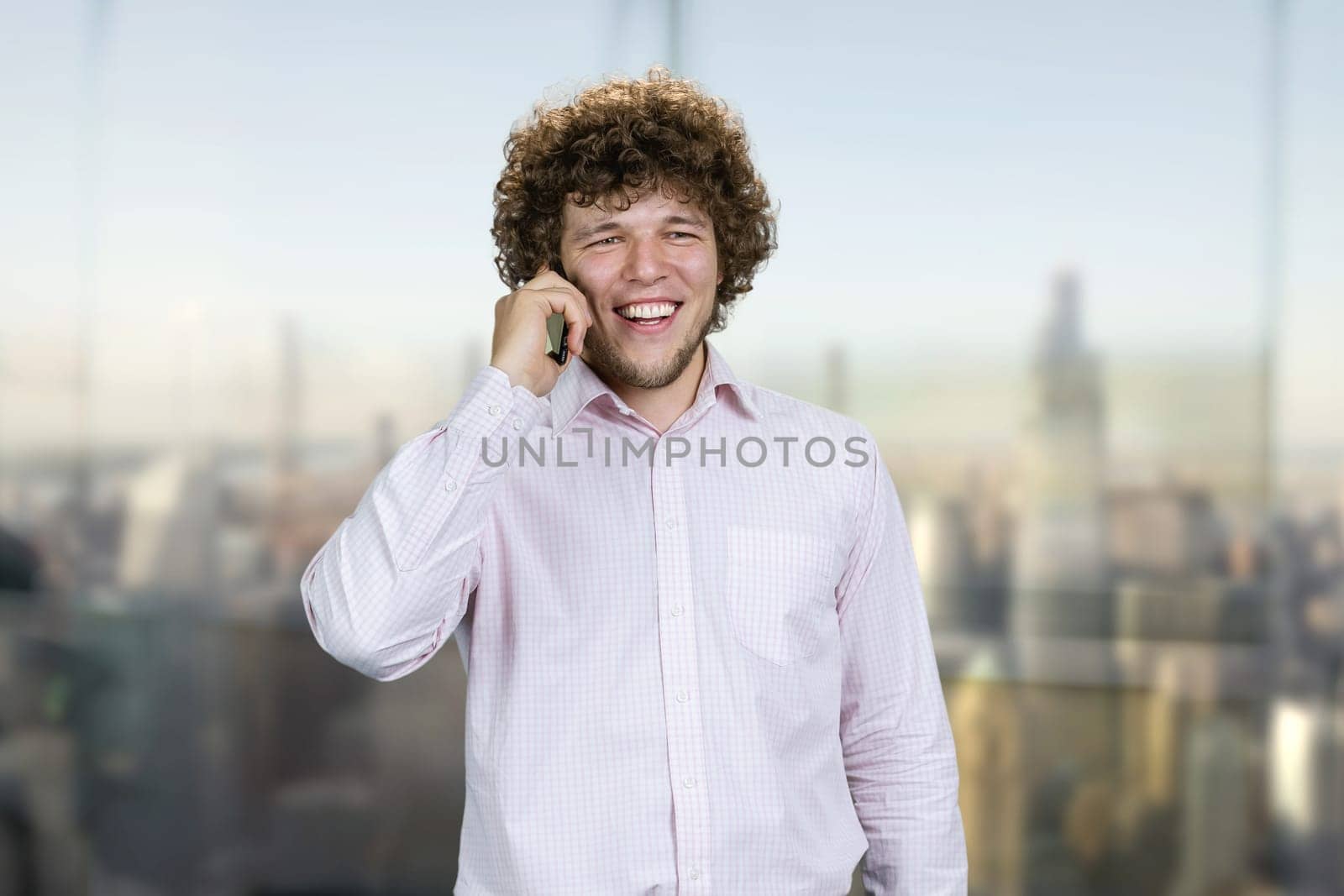Portrait of a young caucasian guy in white shirt talking on the phone. Indoor window with blurred cityscape view in the background.