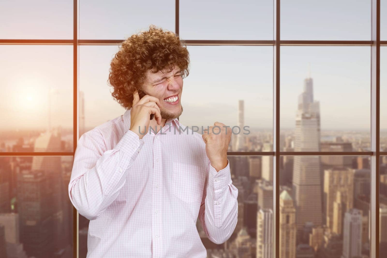 Portrait of a young happy caucasian man talking on the phone rejoicing success. Checkered window with evening cityscape view.