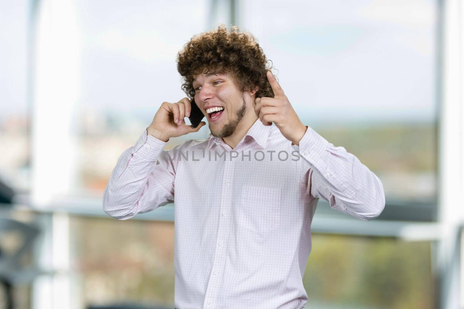 Portrait of a young happy man with curly hair talking on the phone rejoicing success. Indoor window in the background.