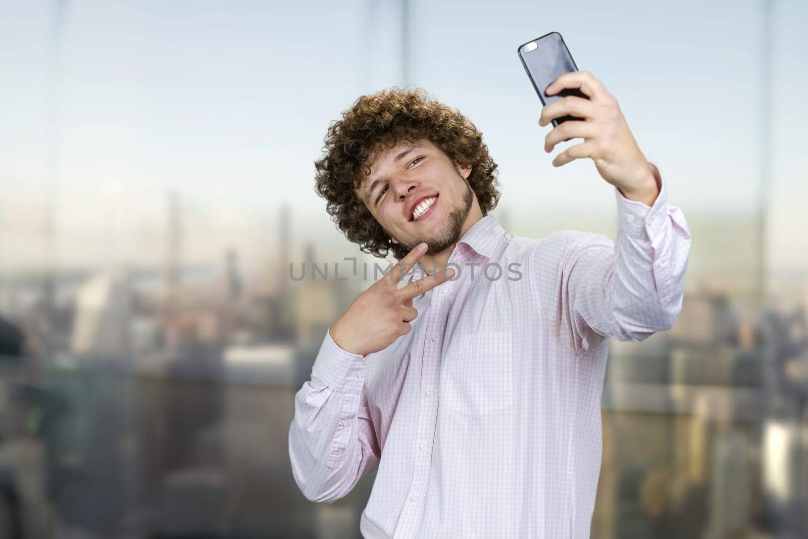 Smiling young man with curly hair making a selfie with victory gesture sign. Urban cityscape in the background.