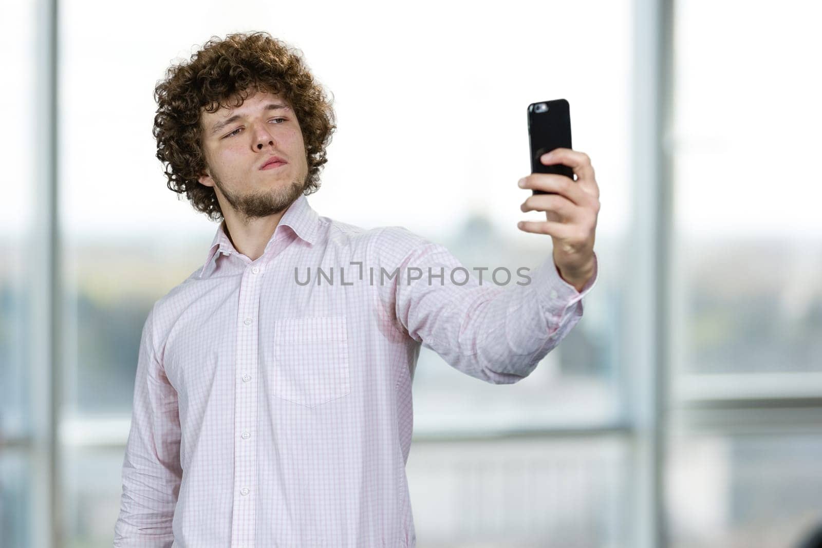 Young man with curly hair making a selfie with arrogant facial expression. Indoor window in the background.