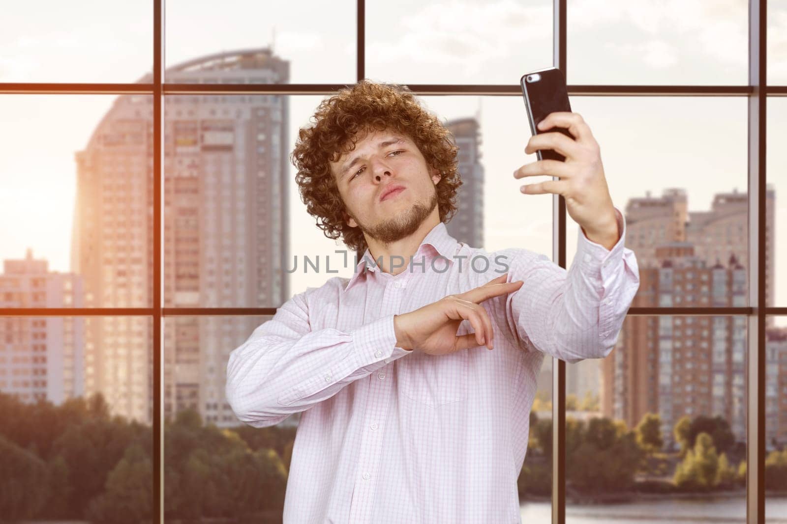 Serious young man with curly hair making a selfie with victory gesture sign. Indoor window with cityscape in the background.