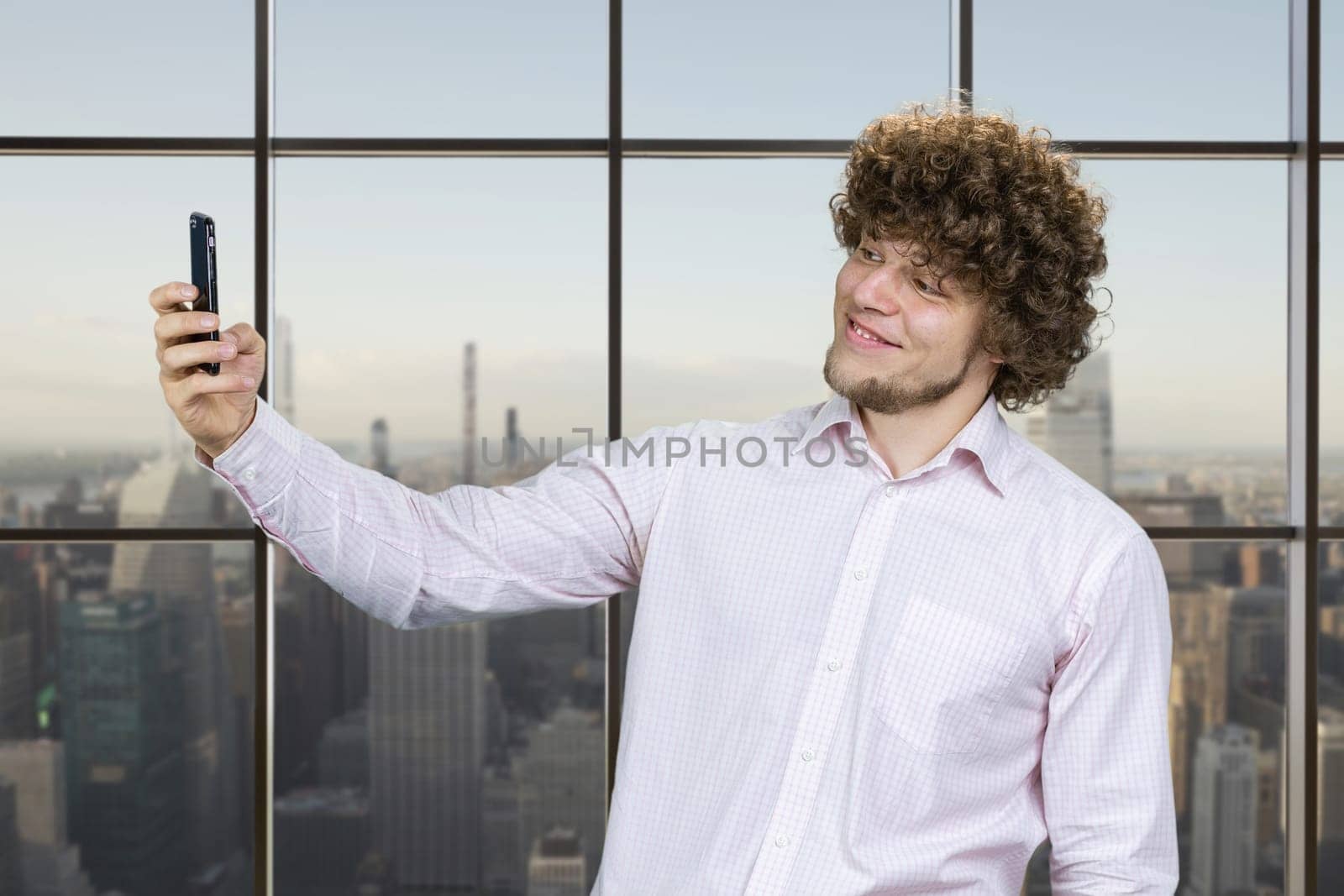 Young man with curly hair making a selfie and smiling with teeth. Indoor window with cityscape in the background.