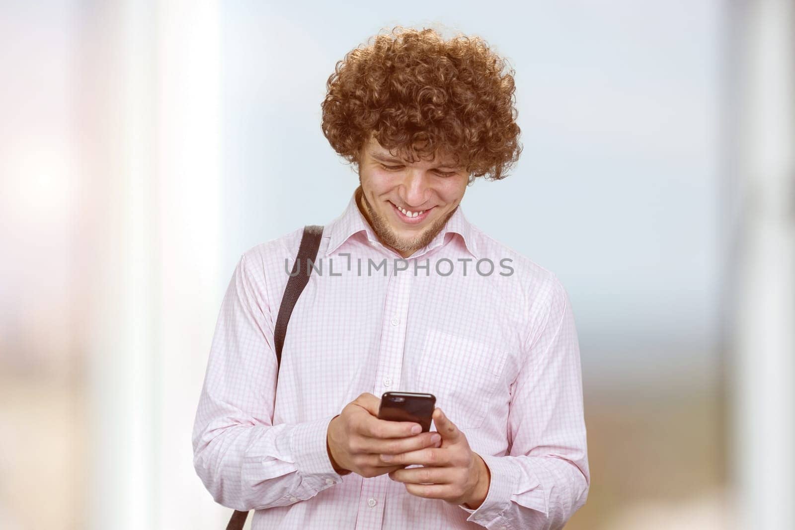 Portrait of a young smiling man with curly hair using his smartphone indoors. Indoor window in the background.
