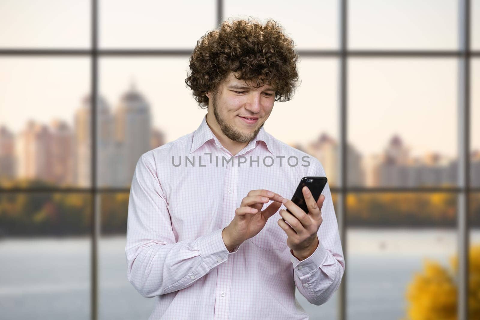 Portrait of a young smiling man with curly hair taps his smartphone. Indoor window with city river view.