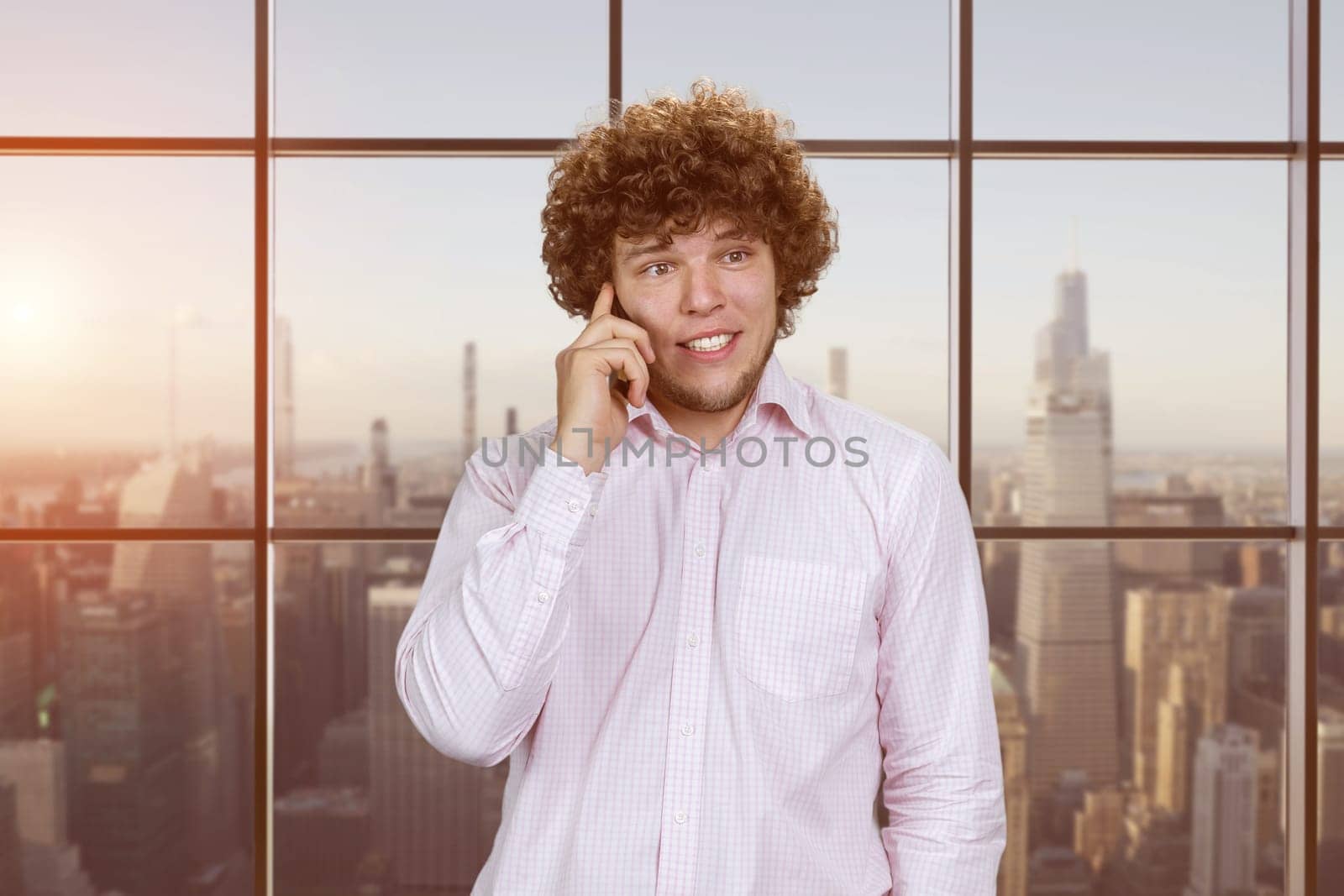 Happy smiling caucasian man in white shirt talking on the phone. Checkered indoor window with evening cityscape view in the background.