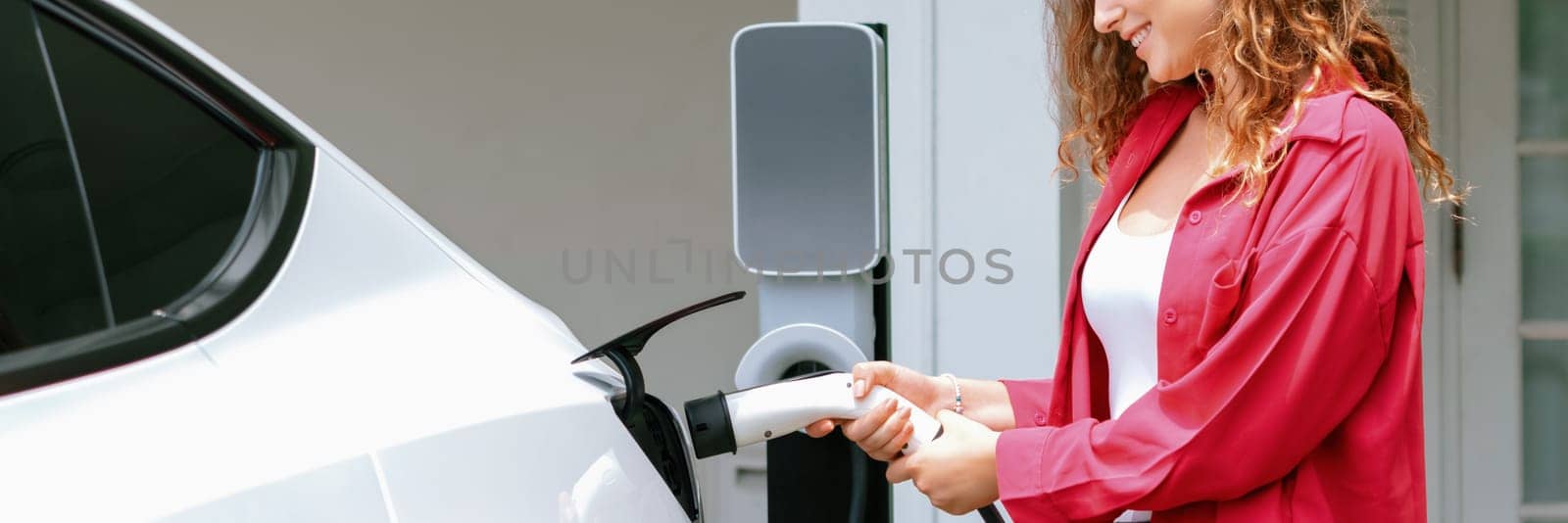 Modern eco-friendly woman recharging electric vehicle from home EV charging station. EV car technology utilized for home resident to future environmental sustainability. Panorama Synchronos