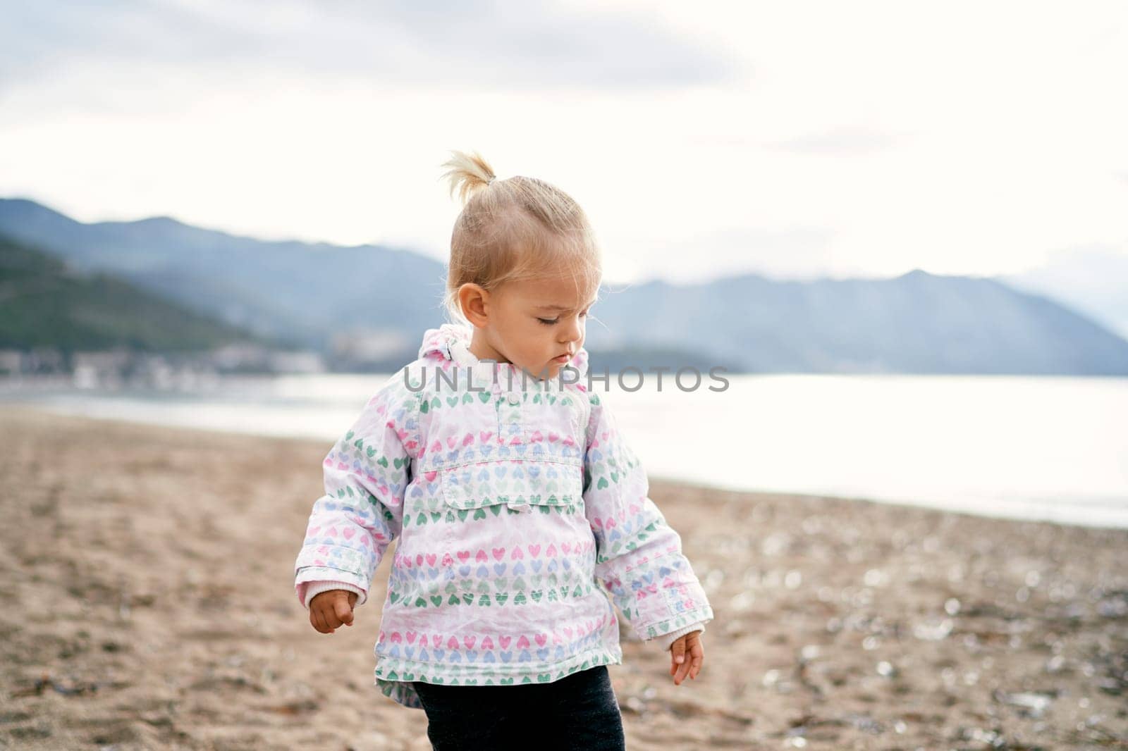 Little girl in a jacket stands on the seashore and looks at her feet. High quality photo
