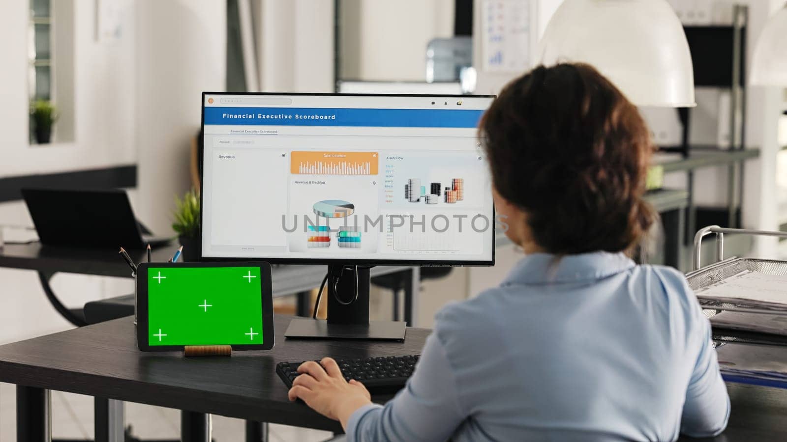 Businesswoman works at desk with greenscreen presented on modern tablet, looking at isolated copyspace template while she analyzes revenue data on pc. Worker uses chromakey display. Handheld shot.