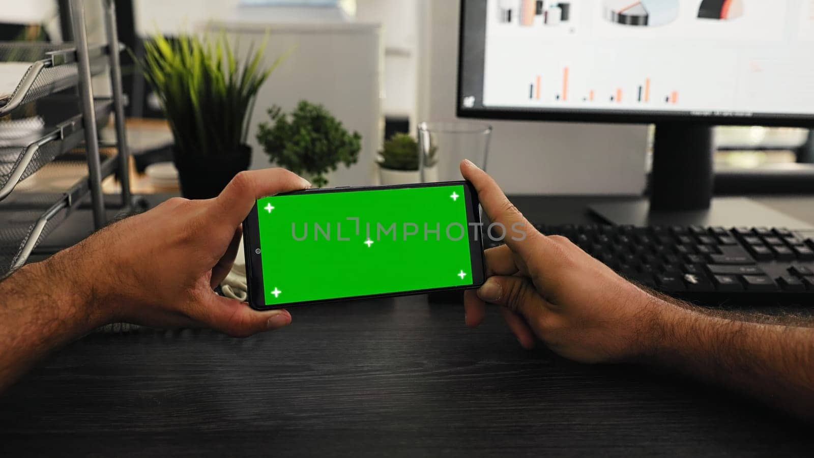 POV of worker holds phone with greenscreen display at desk, working with isolated mockup template at workstation. Young employee looking at mobile phone screen with chromakey.