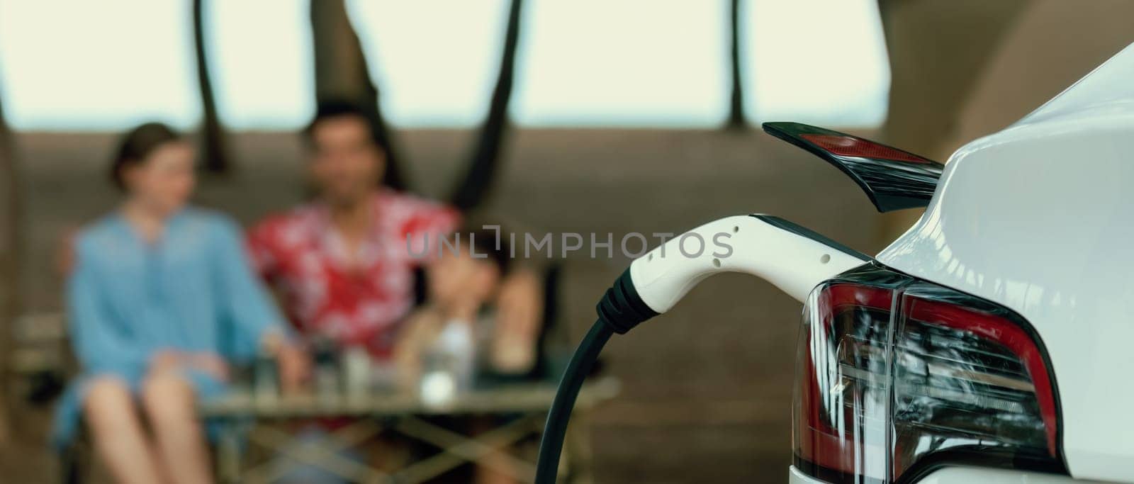 Alternative family camping trip traveling by the beach with electric car recharging battery from EV charging station with blurred family enjoying the seascape campsite background. Perpetual