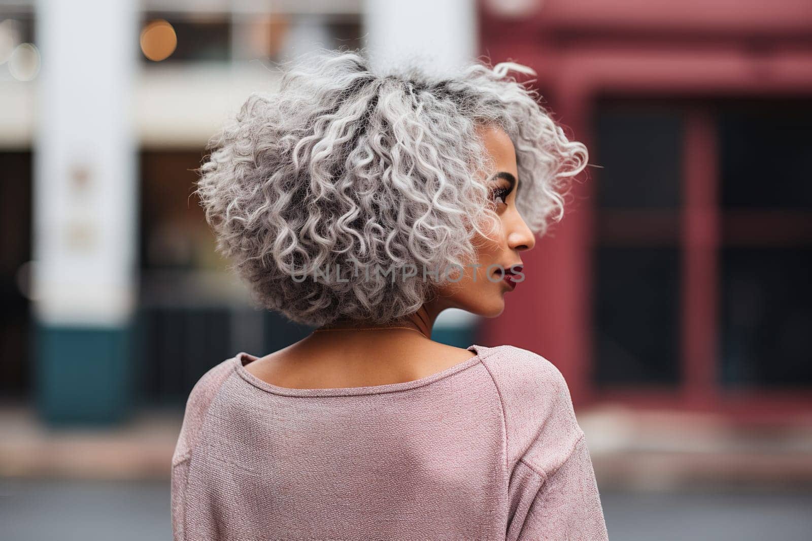 An attractive dark-skinned young woman with curly hair stands backwards in the city.