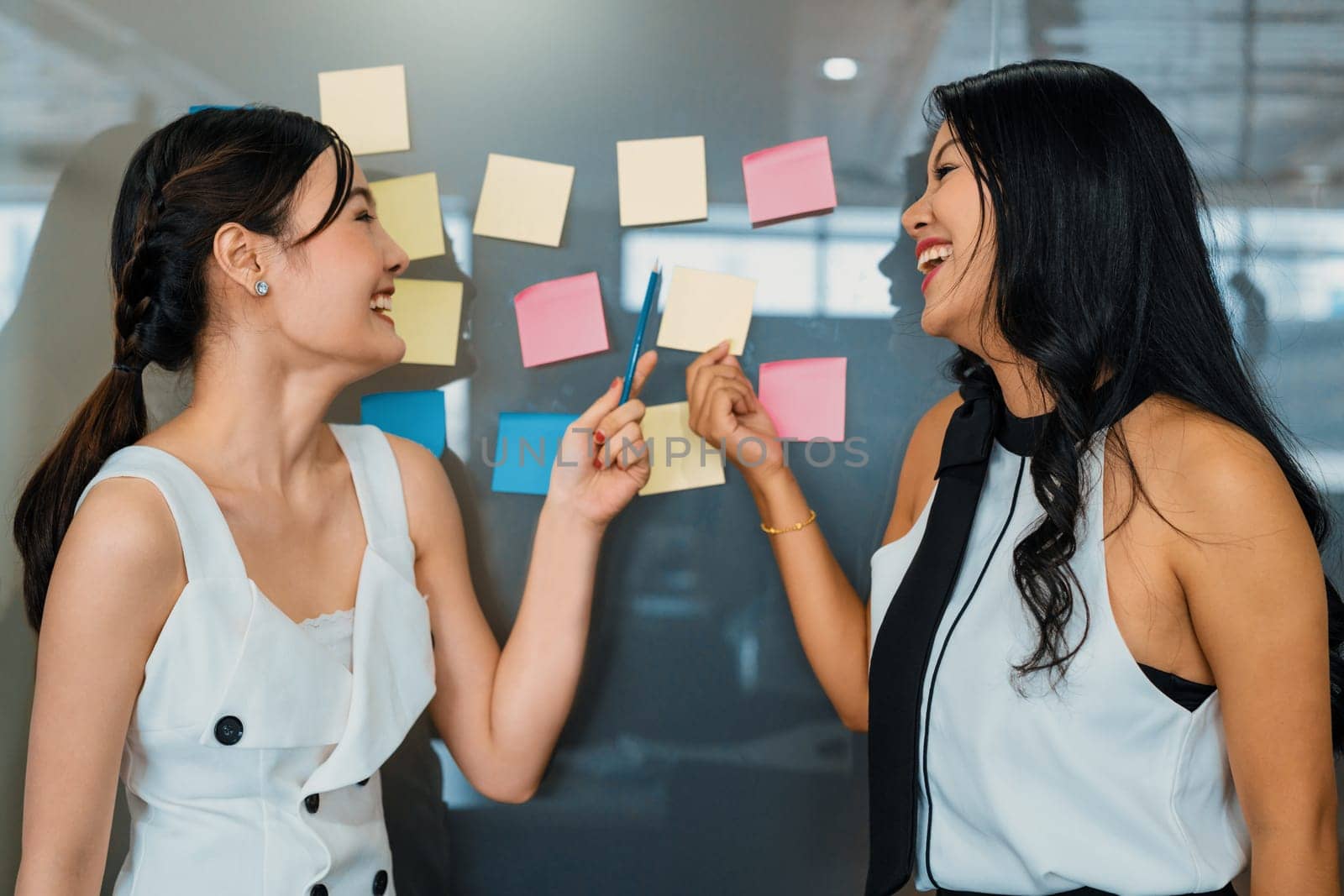 Two young beautiful Asian businesswomen talk and discuss creative post on the window. It is project reminder and thinking idea notes. uds