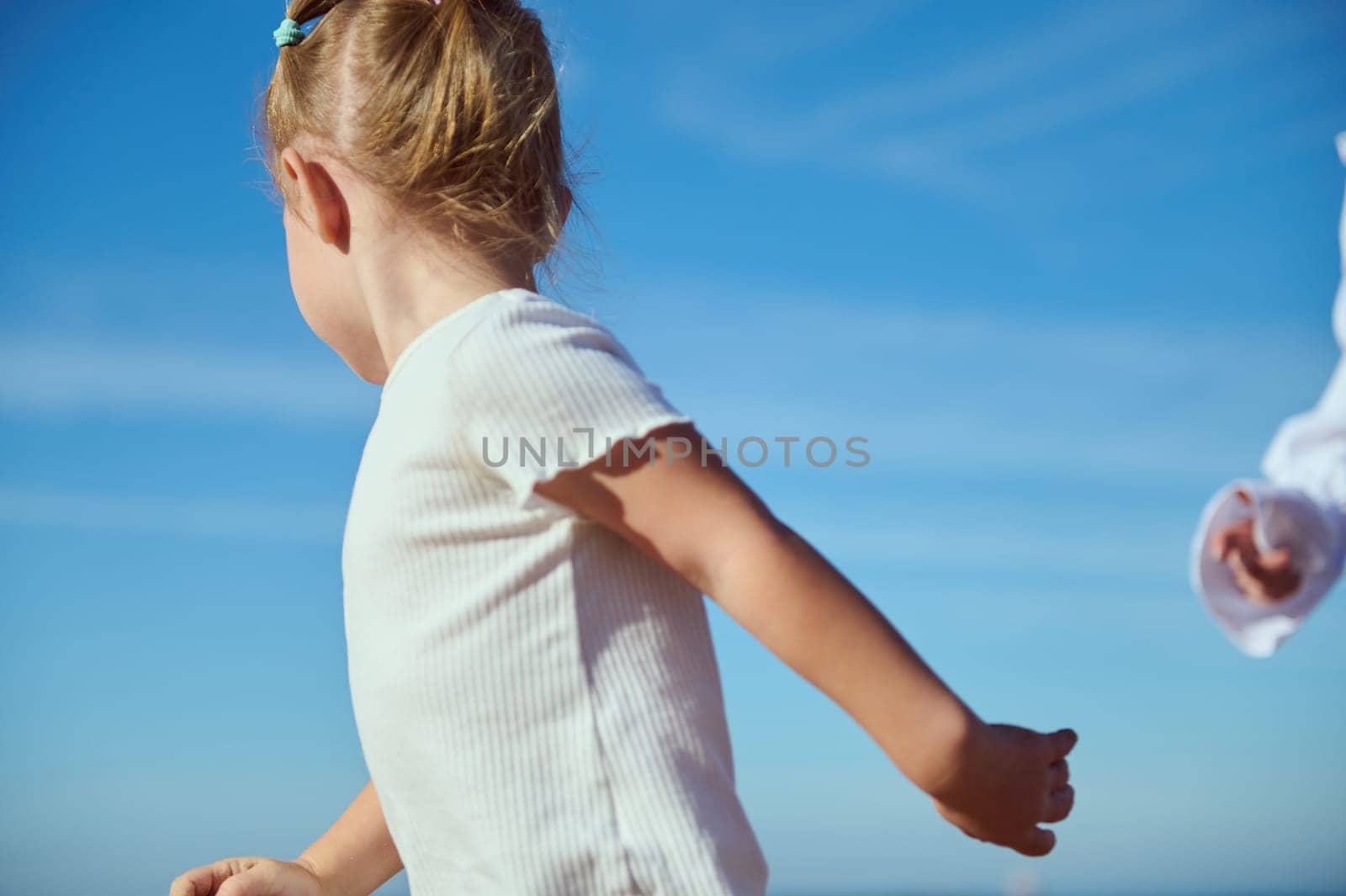 Side portrait of a blonde Caucasian little child girl running against the blue clear sky background. Happy carefree childhood. Kids. Children. People. Family. Lifestyle. Weekend and leisure activity