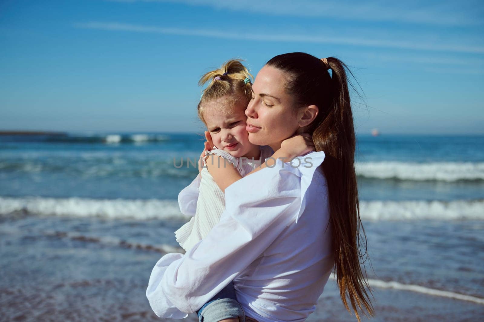Authentic portrait of a beautiful young Caucasian woman carrying and gently hugging her little daughter, enjoying happy time together while walking on the beach. People. Lifestyle. Leisure activity