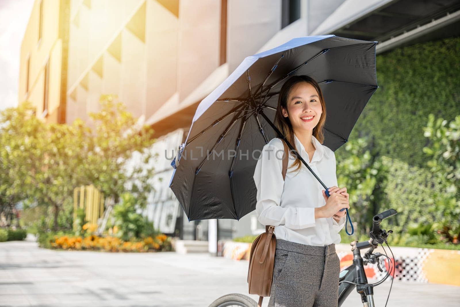 Amid the sunny countryside businesswoman stands with her bicycle and umbrella smiling and carefree. Her playful spirit and fashionable attire blend with beauty of meadow embracing season charm. by Sorapop